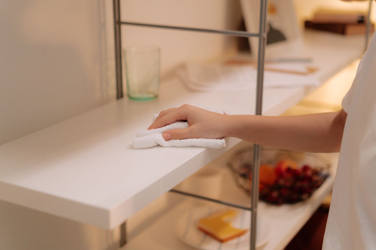 expertise-01 A person cleans a white shelf with a cloth, focusing on home maintenance.